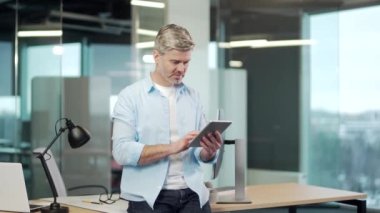 Olgun bir iş adamı modern bir ofiste dijital tablet kullanır. Gri saçlı kıdemli erkek işçi akıllı telefon kullanıyor. İş yerinde günlük gömlekli bir iş adamı çalışıyor.