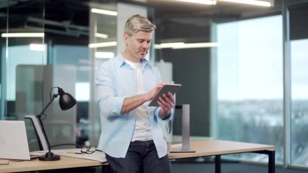 成熟的商人在室内的一个现代化的办公室里使用数字平板电脑 头发灰白的高级男性员工使用浏览智能手机 穿着休闲装在工作场所工作的商人 — 图库视频影像