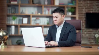Modern bir ofis masasında dizüstü bilgisayarda çalışan düşünceli Asyalı iş adamı portresi. Kendine güveni tam, resmi takım elbiseli dalgın bir adam. Bir sorunu çözmek için ilham almayı düşünüyorum. Asya
