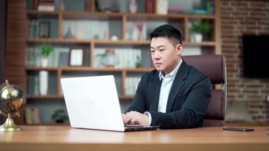Modern bir ofis masasında dizüstü bilgisayarda çalışan düşünceli Asyalı iş adamı portresi. Kendine güveni tam, resmi takım elbiseli dalgın bir adam. Bir sorunu çözmek için ilham almayı düşünüyorum. Asya