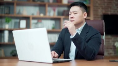 Modern ofis masasında bilgisayar üzerinde çalışan düşünceli Asyalı erkek iş adamı portresi. Düşünceli, düşünceli, takım elbiseli bir iş adamı. Bir sorunu çözmek için ilham almayı düşünmek.