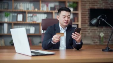 Asian business man shopping online pays by credit card using mobile phone smartphone sitting at desk office, work workplace. Purchase travel, bank service, booking. Male businessman worker in suit