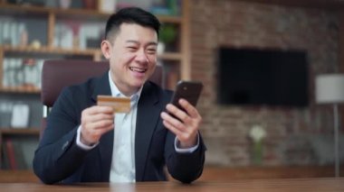 Asyalı iş adamı internet üzerinden cep telefonu akıllı telefonuyla iş yerinde ve masa başında kredi kartıyla alışveriş yapıyor. Satın alma seyahati, banka servisi, rezervasyon. Takım elbiseli erkek işadamı.