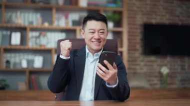 Happy mature Asian business man in office looking at mobile phone with emotion winner or win, financial stock sports betting. Male joyfully exclaims playing game. Excited overjoyed celebrating success