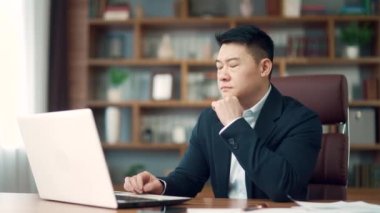 Modern ofis masasında bilgisayar üzerinde çalışan düşünceli Asyalı erkek iş adamı portresi. Düşünceli, düşünceli, takım elbiseli bir iş adamı. Bir sorunu çözmek için ilham almayı düşünmek.