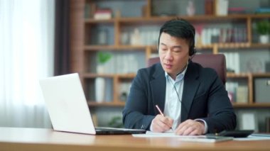 Asian employee studying online virtual tutor using laptop in office. distant worker wears headphones video calling makes notes, elearning with teacher, attending remote conference meeting training
