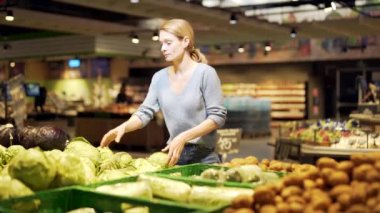 Genç bir kadın markette sebze, lahana ya da meyve seçer, toplar ve inceler. Kadın müşteri tezgahın yanında bir bakkal duruyor ve yiyecek pazarından alışveriş yapıyor.