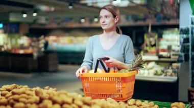 portrait of a young happy female shopper in a grocery store with a basket full of groceries. Smiling woman customer looking at camera while standing in vegetable department of supermarket, market