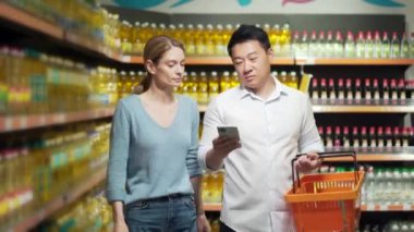 Genç mutlu Asyalı çift süpermarkette akıllı telefon kullanıyor alışveriş arabalarıyla markette ürünleri seçiyorlar. Akıllı telefonları neşeyle tarıyorum. Günlük yiyecek alışveriş listesi. 2 kişi bir seçim yapar.
