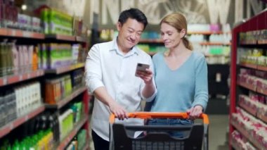 Young happy Asian couple using smartphone in supermarket with shopping cart choosing products while grocery store. browsing smart phone joyful. daily food shopping list. 2 people make a choice
