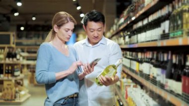 Asian couple chooses wine in a supermarket or grocery store in the alcohol department using a mobile phone smartphone app. husband and wife, man and woman scan the label through use application