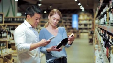 Asian couple chooses wine in a supermarket or grocery store in the alcohol department using a mobile phone smartphone app. husband and wife, man and woman scan the label through use application
