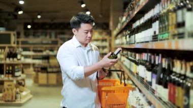 Asian man chooses wine in liquor store or supermarket. The male buyer examines the bottle of wine and puts it in a basket. Male Choice and Buy alcohol from the shelves in the market. 