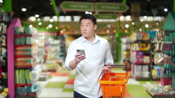 Asian Male Shopper Walks Supermarket Shopping Cart Looking Smart Phone — Video Stock