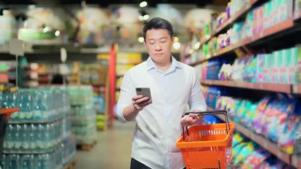 Asian Male Shopper Walks Supermarket Shopping Cart Looking Smart Phone — 图库视频影像