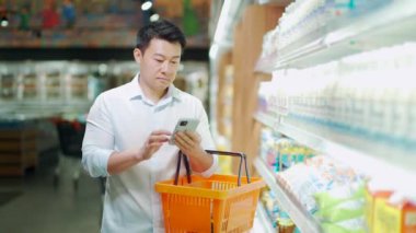 Asian male shopper pick in a grocery store is standing near the supermarket shelves and checking his smartphone. Reads shopping list. use a smart phone. Man customer compares and makes a choice milk