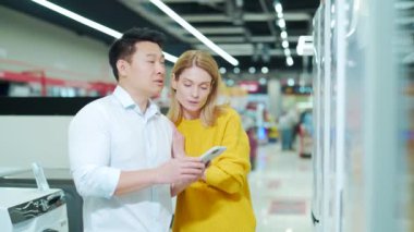 happy mixed race asian couple family man and woman make a choice choose of new household home appliances in supermarket survey Refrigerator Fridge cooler, freezer, store purchase use a smartphone 