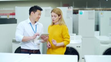 Asyalı erkek danışman, mağazadaki kadın ev aletleri alıcısına profesyonel tavsiyelerde bulunuyor. Asistan müşteriye çamaşır makinesini açıklıyor. Modern elektrik gösteriliyor 