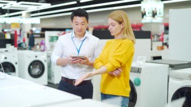 Asyalı erkek danışman, mağazadaki kadın ev aletleri alıcısına profesyonel tavsiyelerde bulunuyor. Asistan müşteriye çamaşır makinesini açıklıyor. Modern elektrik gösteriliyor 