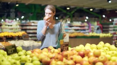 Genç bir kadın süpermarkette sebze ya da meyve seçiyor. Tezgahın yanında bir markette duran kadın müşteri tekrar kullanılabilir bir paket alır ve piyasaya sunar.