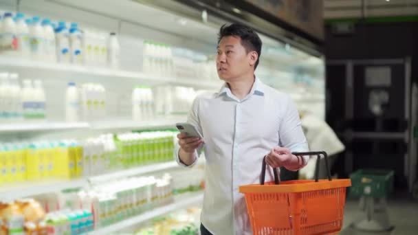 Asian Male Shopper Walks Supermarket Shopping Cart Looking Smart Phone — Stock videók