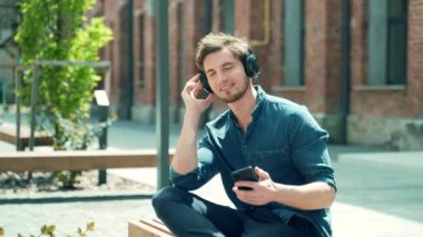 young handsome man sits on a bench and listens hearing music in headphones holding a smartphone in his hands. smiling guy on a city street on urban background. Trendy stylish student relaxes and rests