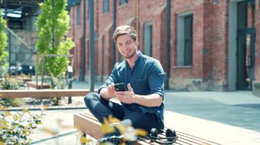 happy young guy student in casual denim shirt using smartphone while sitting on bench in campus. Trendy handsome man browsing chatting text message use smart mobile phone on urban city street. outside