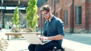 Trendy handsome caucasian man using laptop outdoors, working online in urban city area on a bench. young male student freelance guy in denim casual shirt and glasses uses a computer in campus outside