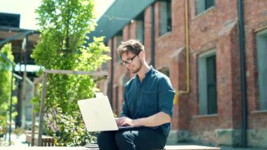 Trendy handsome caucasian man using laptop outdoors, working online in urban city area on a bench. young male student freelance guy in denim casual shirt and glasses uses a computer in campus outside