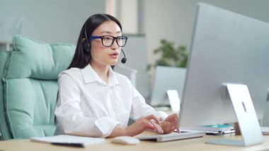 Corporate operator working in customer support service on helpline telesales Focused asi woman representative call center agent in wireless headset helping client complaints using computer in office