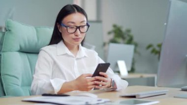 Mutlu Asyalı iş kadını girişimci ofiste otururken akıllı telefon kullanıyor. Bilgisayar masasında cep telefonu kullanan, gezen, sohbet eden, cep telefonlu mesajlar atan genç bir bayan. Gülümse.