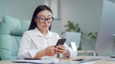 Mutlu Asyalı iş kadını girişimci ofiste otururken akıllı telefon kullanıyor. Bilgisayar masasında cep telefonu kullanan, gezen, sohbet eden, cep telefonlu mesajlar atan genç bir bayan. Gülümse.
