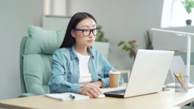 young asian worker, employee entrepreneur studying online remotely using video call remote lessons sitting at desk office Workplace. Woman writes in notebook takes notes while looking at laptop screen