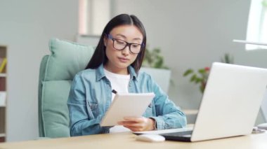 portrait young asian girl student listen studying online using video call remote lessons sitting at desk at home or classroom. indoor. Woman writes in notebook takes notes while looking at camera 