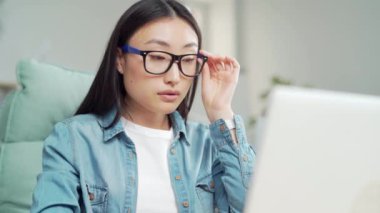 Modern ofiste dizüstü bilgisayarla çalışan gözlüklü ve sıradan giysili güzel Asyalı kadın. İşçi işyerinde masa başında oturuyor. Çalışan bilgisayar taraması ve mesaj kullanıyor. Asya