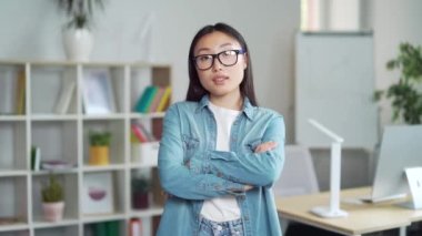 Modern ofiste kollarını kavuşturup kameraya bakan ciddi bir Asyalı kadın ofis çalışanı. Profesyoneller, iş yerinde yaratıcı ve başarılı bir iş kadını.