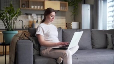 pregnant woman works on a laptop computer online remotely sitting at home on the sofa in living room. jobs during pregnancy on the Internet. Business female employee typing texting browsing chatting