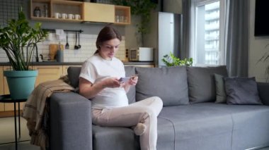 pregnant woman shopping online on the Internet store. Female makes a payment using a mobile phone with a smartphone and uses plastic credit card while sitting at home on the sofa. pays for purchases