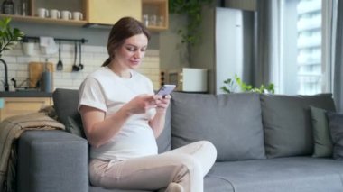 Pregnant Caucasian young woman using mobile phone smartphone at home. Happy female enjoy pregnancy use shopping online store using app browsing chatting texting messaging 