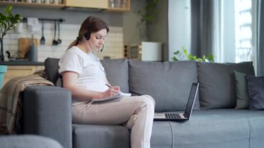 Maternity leave. Pregnant freelancer woman sitting at home on sofa with laptop and in headphones in home clothes. Works on the laptop computer. remote online course, meeting, conference or training
