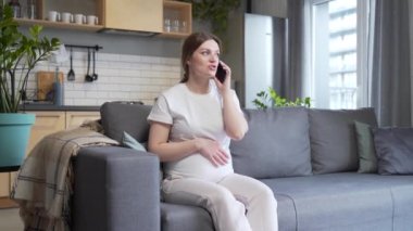 Expectant Female With Smartphone On Sofa. Happy pregnant woman talking on cellphone at home. mother sitting on couch, speaking on mobile phone, touching belly. Consulting