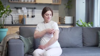 Young expectant pregnant woman, vaccinated against the virus, looks at the camera and smiles, sits at home on the couch, and points his finger at the patch and the injection site on the shoulder
