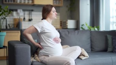 young pregnant woman with back pain sitting on the sofa and massaging her lower back at home on the sofa. The female suffers discomfort holding the waist during pregnancy difficulties backache