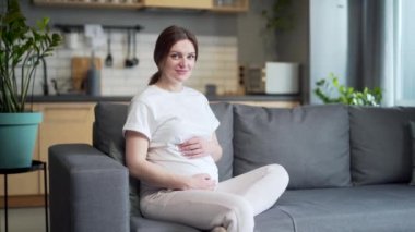 portrait young happy pregnant Caucasian attractive woman sitting on sofa at home stroking belly looking at camera. Smiling cheerful female during pregnancy Happiness is expecting a baby in room 