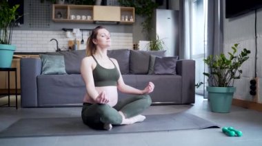 young active pregnant woman doing yoga sitting on mat in lotus pose indoors in living room. at home Practicing yoga during pregnancy. Meditation and relaxation