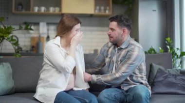 despair and mood swings in a pregnant woman. female upset crying during pregnancy. The couple is sitting on the sofa at home, the husband supports and calms the wife. bad mood and irritation in woman