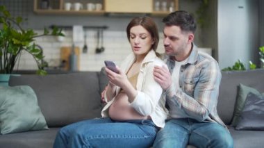 happy young married couple sitting on sofa at home choosing for future unborn child. pregnant wife and husband make purchases online using a mobile phone smartphone together shop Internet store