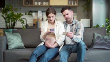 Expectant married couple pregnant wife and husband shopping online paying bills using mobile phone smartphone and credit card sitting at home on sofa. Family during pregnancy, buying goods in store