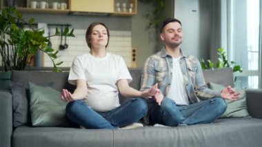 young happy couple practicing yoga during pregnancy sitting on sofa in lotus position. pregnant wife and husband spend time together, relax and meditate at home. Happiness, love and support in family