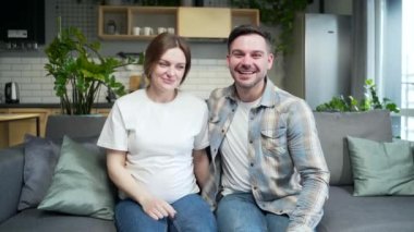 young happy couple chatting online via video call. Pregnant wife and husband show heart love sign together look at camera. Sitting on sofa at home. Remote meeting with friends or family. View webcam